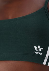 Žalias sportinis liemenėlių, pasižymintis patogiu prigludimu, pagamintas iš minkštos medžiagos, su baltu Adidas logotipu ir juostelėmis šonuose.