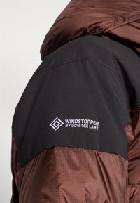 Giacca isolante marrone con una texture liscia e lucida. Patta nera sulla spalla con il logo bianco "WINDSTOPPER by GORE-TEX LABS".