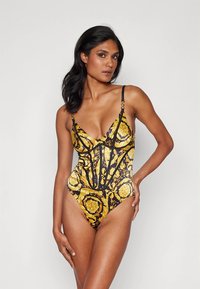 Juoda ir auksinė vienspalvė maudymosi kostiumėlis su gilia V formos iškirpte, dekoratyvinėmis siūlėmis ir reguliuojamomis petnešėlėmis. Puošia išskirtiniai aukso raštai.