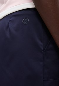 Gros plan sur une personne portant un pantalon bleu marine avec un logo circulaire noir comportant la lettre "B" près de la taille et une poche latérale.