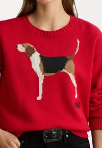 Rød strikket genser med et beagle-design i nyanser av brunt, svart og hvitt; ribbet rund hals og mansjetter; små broderte initialer på kanten.