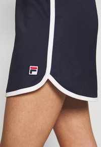 Jupe bleu marine avec un ourlet incurvé, un bord blanc et un petit logo brodé avec des accents rouges et blancs près du bord inférieur.