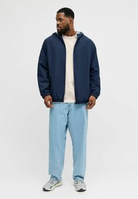 Temno modra jakna s kapuco, elastičnimi manšetami in zadrgo čez svetlo bež majico; kombinirana s svetlo modrimi kavbojkami in sivimi supergami.