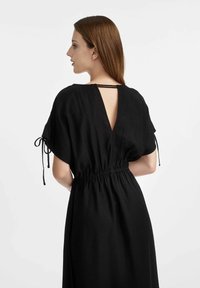 Czarna sukienka z dekoltem w kształcie litery V z tyłu i elastycznym pasem, z krótkimi rękawami z detalami wiązania, wykonana z lekkiego materiału o gładkiej teksturze.