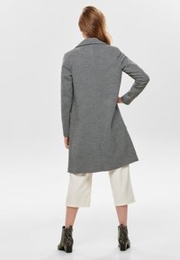 Donna in piedi con una mano sul fianco, indossa un cappotto grigio di lunghezza media, pantaloni bianchi corti e stivaletti in pelle di serpente.