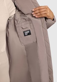 Lysbrun, quiltet pufferjakke indvendig med en sidelomme. Sort label med hvid tekst syet på lommen. Glat, skinnende stof.