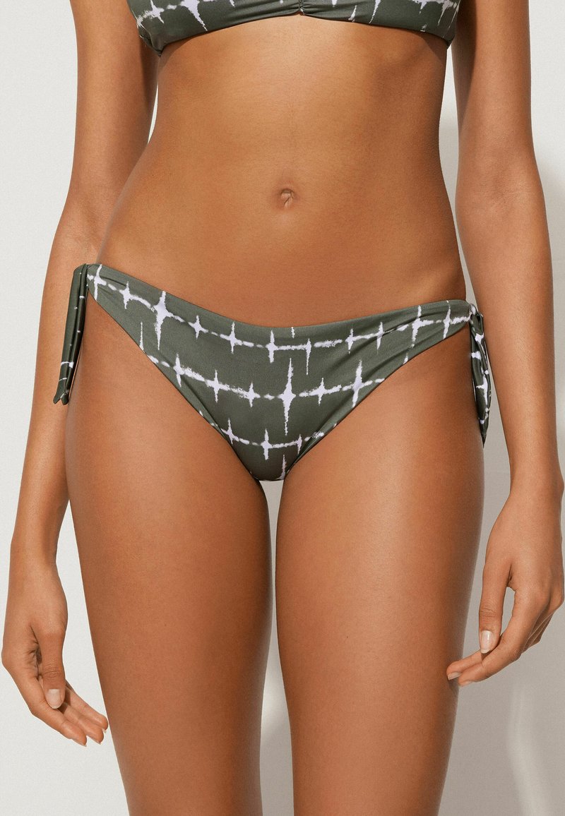 Bas de bikini vert avec un motif géométrique blanc, des accents à nouer sur les côtés et une texture lisse et extensible.