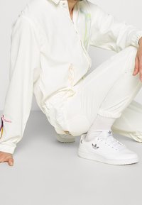 Persoon in een witte casual outfit met kleurrijke lijntekeningen op de mouwen en witte Adidas sneakers, hurkend op een lichte vloer.