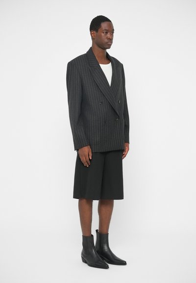 Dwurzędowa czarna marynarka w paski z dużymi klapami, połączona z dopasowanymi czarnymi szortami i czarnymi skórzanymi botkami za kostkę.