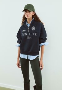 Námornícky sveter s grafikou "NEW YORK", biely text, vysoký golier, nosený cez svetlo modrú pruhovanú košeľu, skombinovaný so zelenými legínami a čiernymi čižmami.