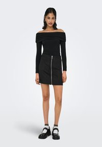 Svart av-shoulder topp med teksturert stoff, kombinert med et svart miniskjørt med glidelås foran. Modellen har på seg svarte sko og hvite sokker.