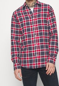 Camicia a quadri rossi e blu con chiusura a bottoni, maniche lunghe e colletto classico. Realizzata in cotone con una consistenza morbida e delicati accenti bianchi.