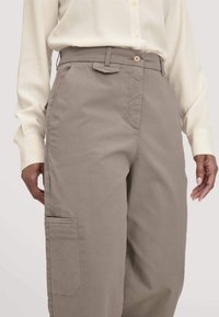 Vrouw in taupe cargobroek en een crèmekleurig overhemd met lange mouwen en knopen, handen ontspannen langs de zij, tegen een effen achtergrond.