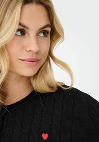 Maglione nero a maglia con motivi a trecce, colletto rotondo e un grande cuore rosso in evidenza sul lato sinistro. Texture morbida e vestibilità rilassata.