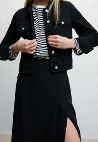 Giubbotto di denim nero con bottoni a pressione argentati, indossato su una maglietta a righe bianco e nera. L'outfit include una gonna nera con uno spacco laterale.