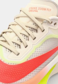 Nike Zoom Fly sneaker in lichtbeige, met een bovenwerk van mesh, neon groene en roze accenten, gestructureerde zool en platte veters.