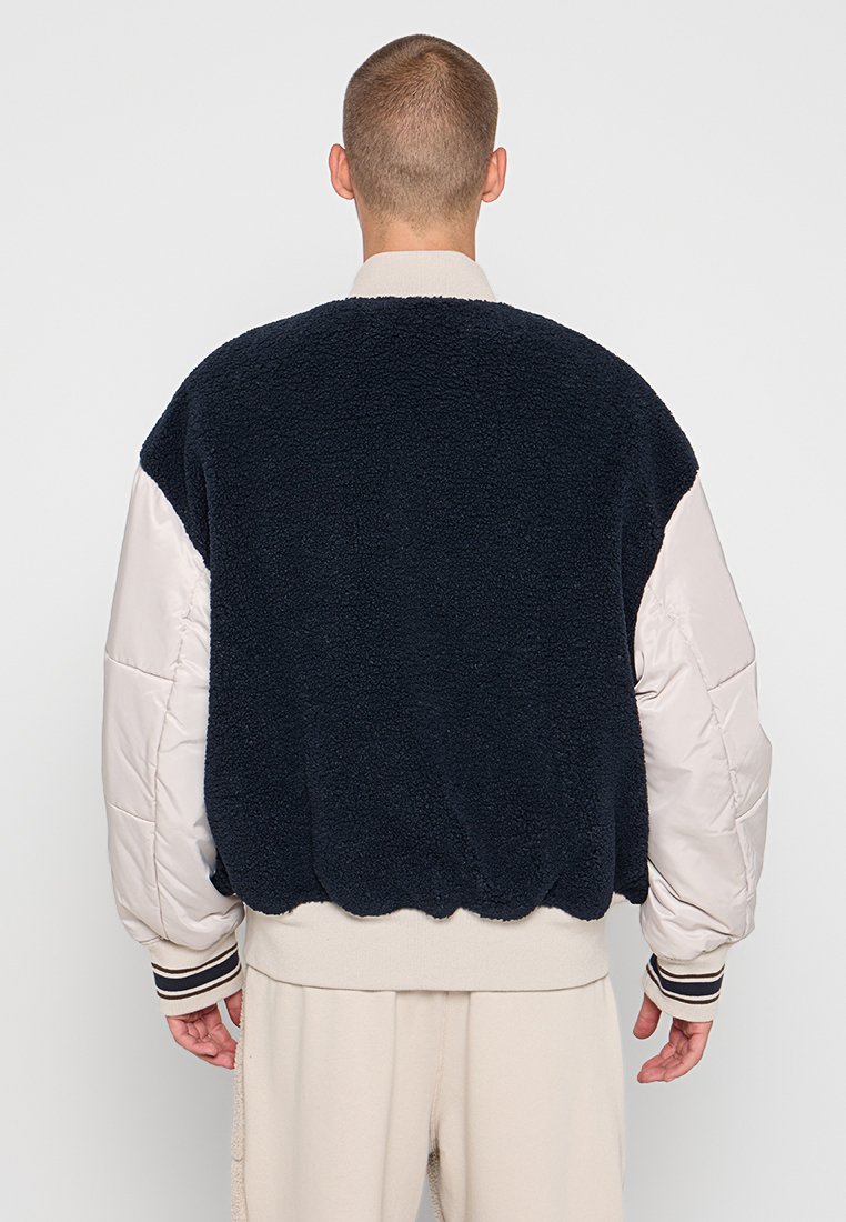 Giacca bomber in pile navy con maniche imbottite crema, polsini a costine e orlo elasticizzato, caratterizzata da una texture morbida e una forma squadrata.