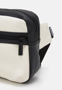 Borsa a tracolla compatta in pelle nera e bianca con zip visibile e etichetta del marchio sul manico, su sfondo bianco.