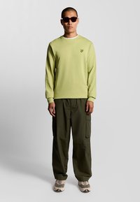 Felpa verde chiaro con collo rotondo e logo ricamato, abbinata a pantaloni cargo oliva e sneaker beige. Il modello indossa occhiali da sole scuri.