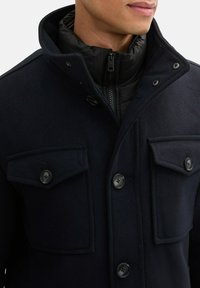 Granatowa kurtka z wełny z domieszką, z wysokim kołnierzem, zamkiem błyskawicznym wewnątrz oraz dwiema kieszeniami na piersi zapinanymi na guziki z płaskimi klapami. Teksturowane wykończenie.