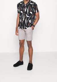 Zwart T-shirt met korte mouwen en abstracte, multicolor patronen, gecombineerd met lichtbeige shorts. Zwarte suède schoenen maken de outfit compleet.