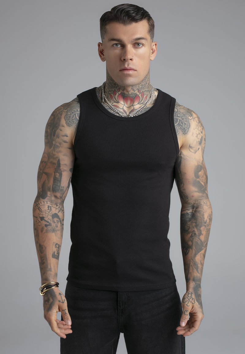 Man met tatoeages op zijn hele arm en nek, gekleed in een zwart mouwloos shirt, zilveren kettingen en armbanden, staand tegen een effen grijze achtergrond.