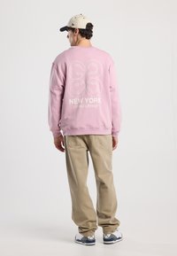 Felpa rosa mostrata da dietro, con un design grafico bianco. Abbinata a pantaloni beige dalla vestibilità ampia e sneaker.