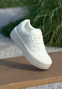 Balti odiniai sportbačiai su lygia pada, apvalia nosimi ir perforuotomis detalėmis. Savybės: raišteliai ir minimalistinis dizainas, padėtas ant medinės paviršiaus.