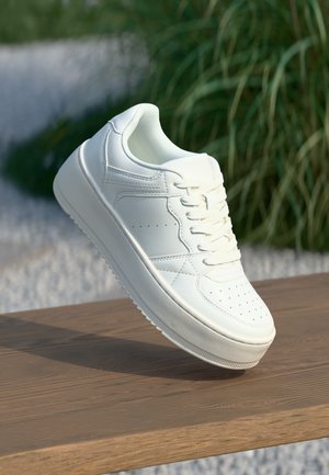 Adidași din piele albă cu talpă plată, vârf rotund și accente perforate. Prezintă un sistem de închidere cu șireturi și un design minimalist, așezați pe o suprafață din lemn.