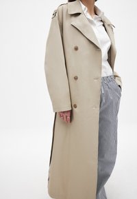 Beidža trenča mēteli ar dubulto pogu dizainu, lielu apkakli un pogu akcentiem. Pāris ar svītrotiem biksēm, radot slāņainu izskatu.