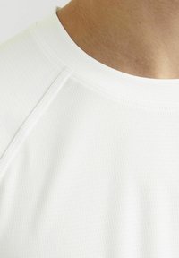 Valge sportlik särk tekstuuriga, hingavast kangast; sellel on ümar kaelus ja lühikesed raglanvarrukad. Minimalistlik õmbluse detail on nähtav.