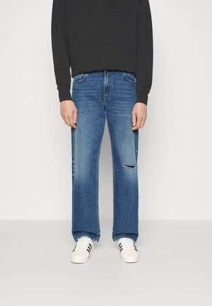Sudadera negra con capucha, vaqueros de denim azul de ajuste relajado, lavado desgastado, detalle roto en la pierna izquierda, y zapatillas blancas con rayas negras.