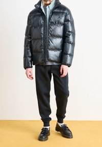 Chaqueta acolchada negra y brillante con patrón de acolchado; cierre de cremallera en el frente; llevada sobre una camiseta gris y pantalones de chándal negros; mocasines negros con detalles de flecos.
