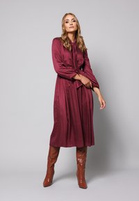 Długa bordowa sukienka w białe kropki, materiał o gładkiej fakturze, z długimi rękawami i wiązanym w talii pasem. Połączona z wysokimi, błyszczącymi brązowymi botkami.