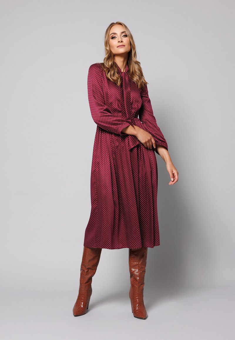 Długa bordowa sukienka w białe kropki, materiał o gładkiej fakturze, z długimi rękawami i wiązanym w talii pasem. Połączona z wysokimi, błyszczącymi brązowymi botkami.
