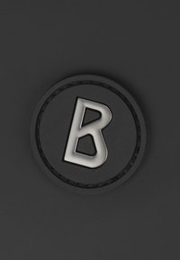 Écusson noir rond présentant un "B" argenté en gras, avec une bordure texturée et une surface intérieure lisse.