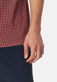 Kurzarmhemd mit einem rot-weißen geometrischen Muster aus strukturiertem Stoff, kombiniert mit blauen Shorts. Detail einer Hand am Rand.