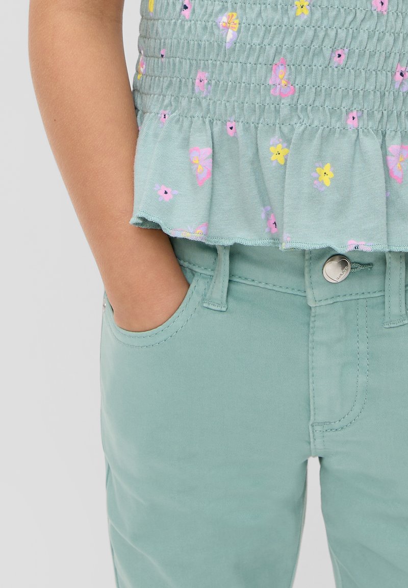 Jasnozielona bluzka z krótkim rękawem z kwiatowymi i motylkowymi wzorami; zestawiona z dopasowanymi zielonymi spodniami. Gładki materiał, elastyczny pas i detal z falbaną.