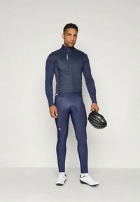 Ναυτικό ποδηλατικό μπουφάν με υφή, συνδυασμένο με εφαρμοστά ναυτικά κολάν. Το άτομο κρατά ένα μαύρο κράνος και φορά λευκά αθλητικά παπούτσια.