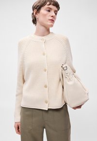 Κρεμ χρώματος πλέκτο καπέλο με στρογγυλή λαιμόκοψη, μπροστινά κουμπιά και υφαντό ύφασμα, συνδυασμένο με μια ανοιχτή μπεζ τσάντα και ελιόχρωμο παντελόνι.