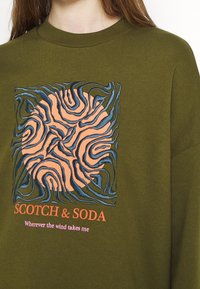 Olive zielony sweter z centralnym abstrakcyjnym wzorem w kolorze pomarańczowym i niebieskim. Napis brzmi "SCOTCH & SODA" oraz "Dokąd zawieje mnie wiatr."