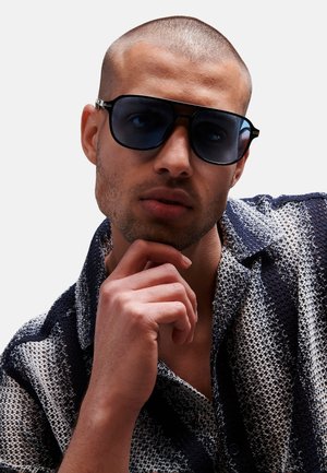 Gafas de sol negras con lentes oscuros, llevadas por un modelo. Él está en una camiseta de punto a rayas azul marino y blanca, apoyando su chin en la mano.