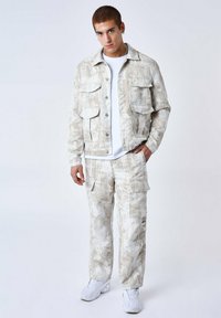 Chaqueta de mezclilla color beige claro con un patrón texturizado, cuatro bolsillos frontales, combinada con pantalones cargo del mismo color. Camiseta blanca por debajo.