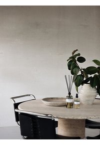 Tavolo rotondo in pietra con superficie texturizzata, sedie in pelle nera con accenti in cromo, vaso in ceramica bianca e diffusori con canne slanciate.