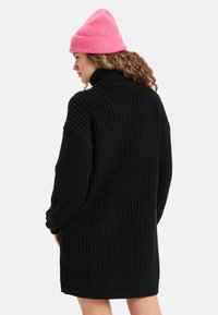 Robe en maille côtelée noire avec un col montant, associée à un bonnet en maille côtelée rose vif, mettant en avant une coupe décontractée et un tissu texturé.