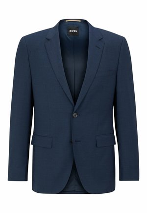 Homme portant un costume bleu ajusté avec une chemise bleu clair, debout devant un fond uni gris clair.