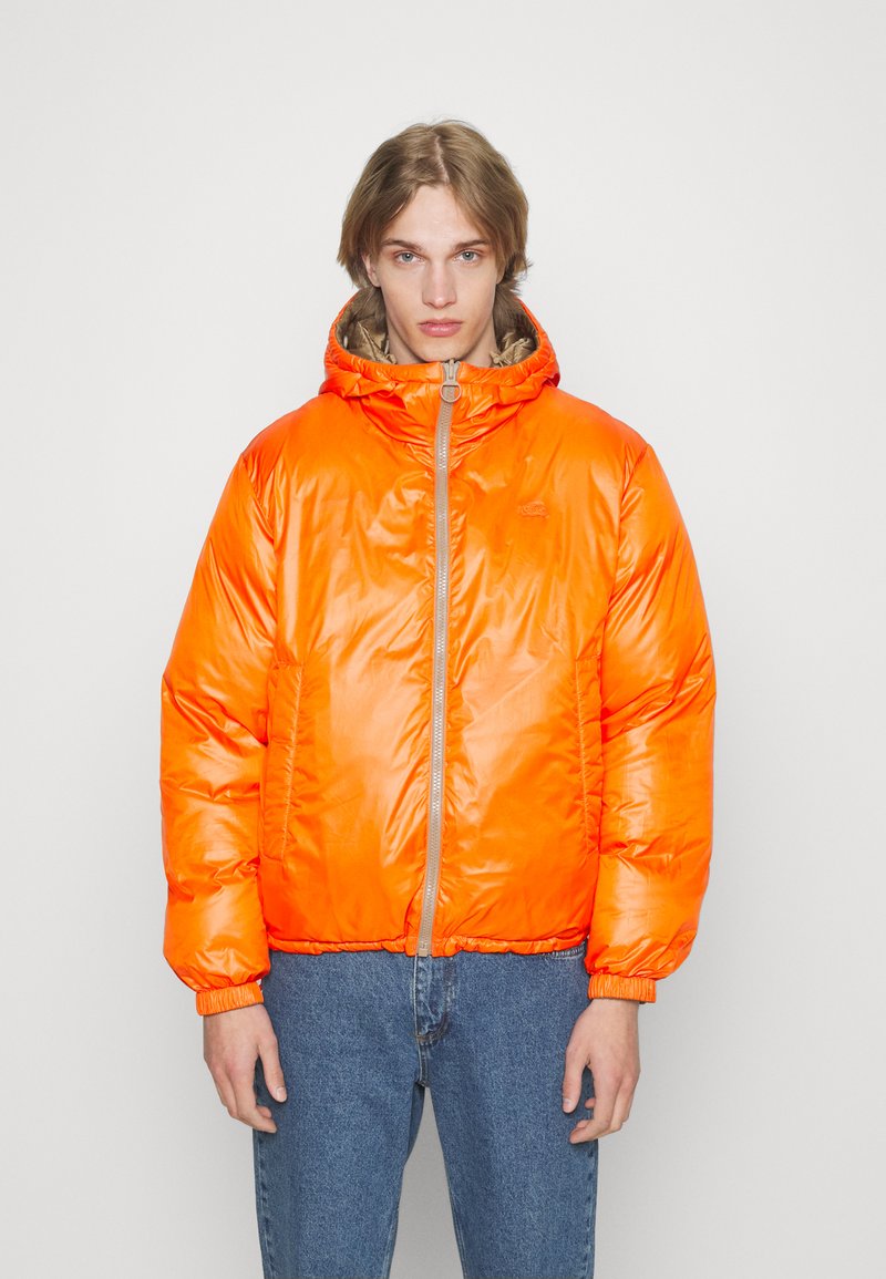 Helle orange Puffjacke mit Kapuze, Reißverschluss und elastischen Bündchen. Aus glänzendem Material und mit lässigem Schnitt. Über blauen Jeans getragen.