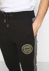 Zwarte joggers met een geribde tailleband, trekkoord, zijzak met rits en een gouden geborduurd logo op de dij. De stof lijkt zacht en glad te zijn.