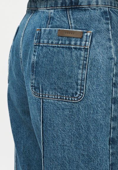 Πίσω όψη μπλε τζιν με τετράγωνη πίσω τσέπη και μια μικρή καφέ ετικέτα με την επιγραφή «soeur» πάνω από την τσέπη.