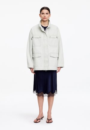 Kvinde iført en lys grå oversized jakke over en mørkeblå knælængde kjole med blonder, kombineret med brune hælede sandaler, stående oprejst.